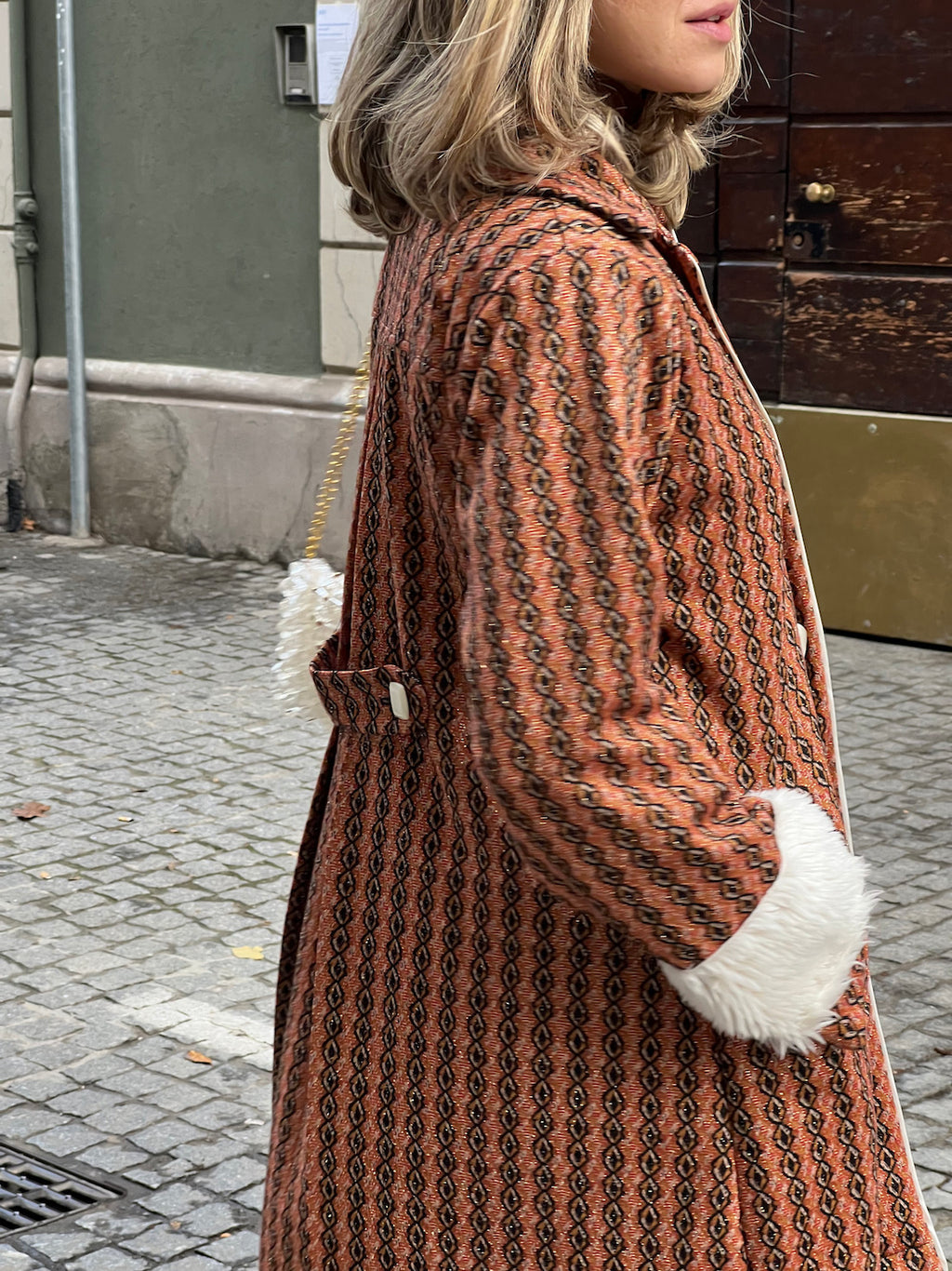 Side view of handmade burnt-orange vintage-inspired coat with geometric pattern, white faux-fur cuffs and buckle-belt detail.
