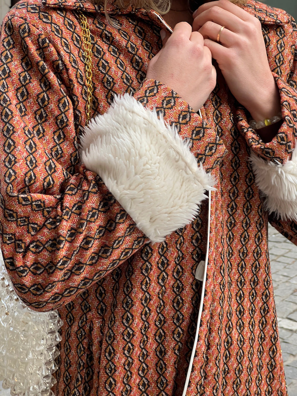 Close-up of handmade burnt-orange vintage-inspired coat with geometric pattern, white faux-fur cuffs, and button closures.